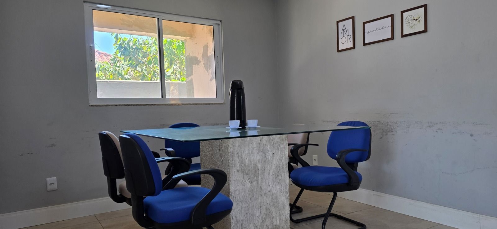 Uma sala com uma mesa de tampo de vidro, cadeiras de escritório azuis e obras de arte emolduradas na parede cinza. Uma janela oferece vista para o exterior.