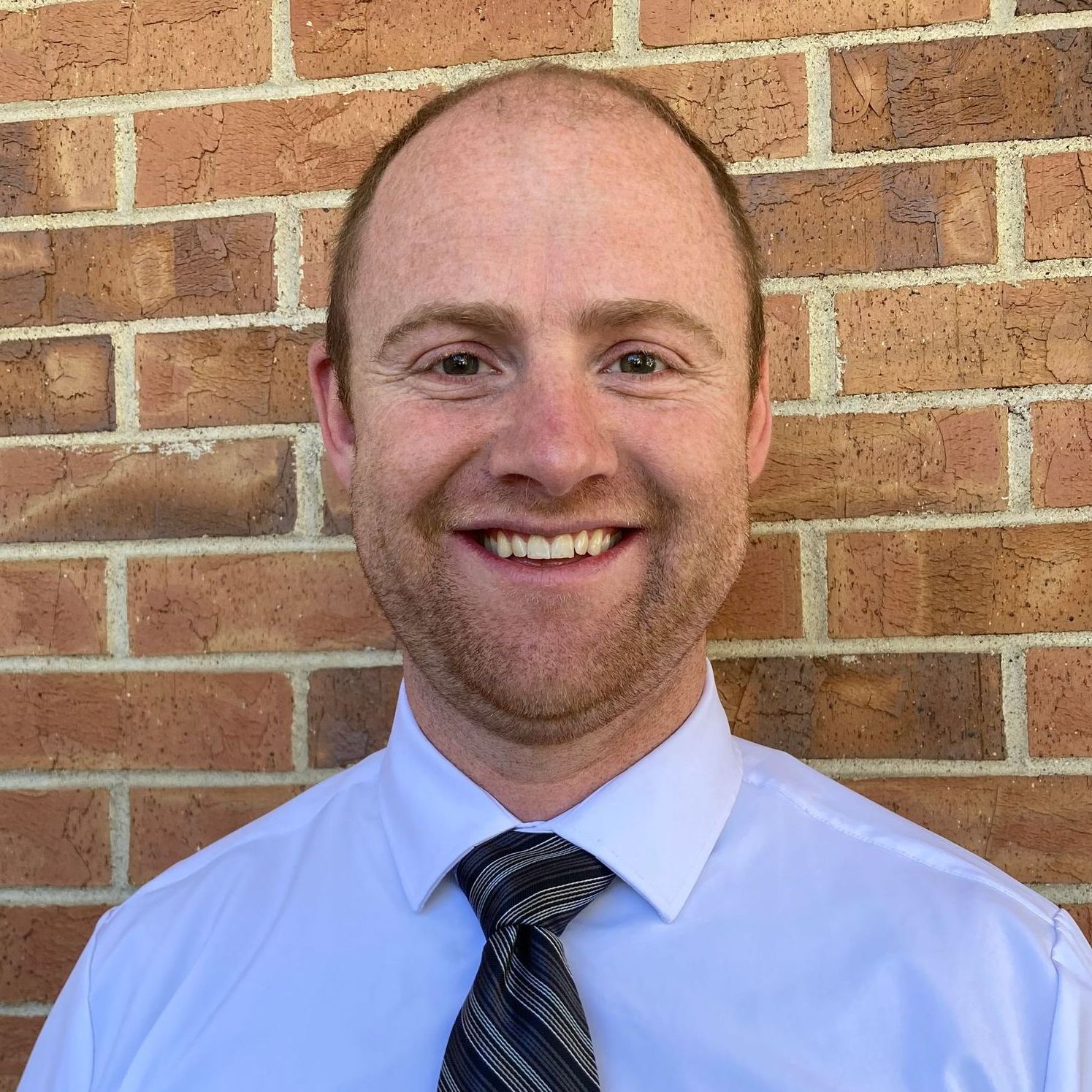 A man in a white shirt and tie is smiling in front of a brick wall.