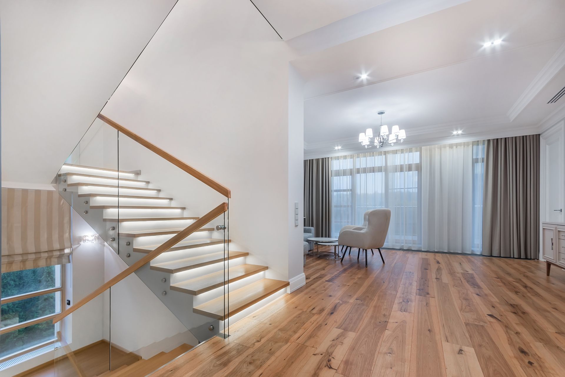 Modern home interior with wooden floors, staircase with glass railing, and a living area.
