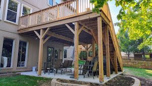 Two-story wooden deck with stairs and support structure over a patio. Green yard and house in background.