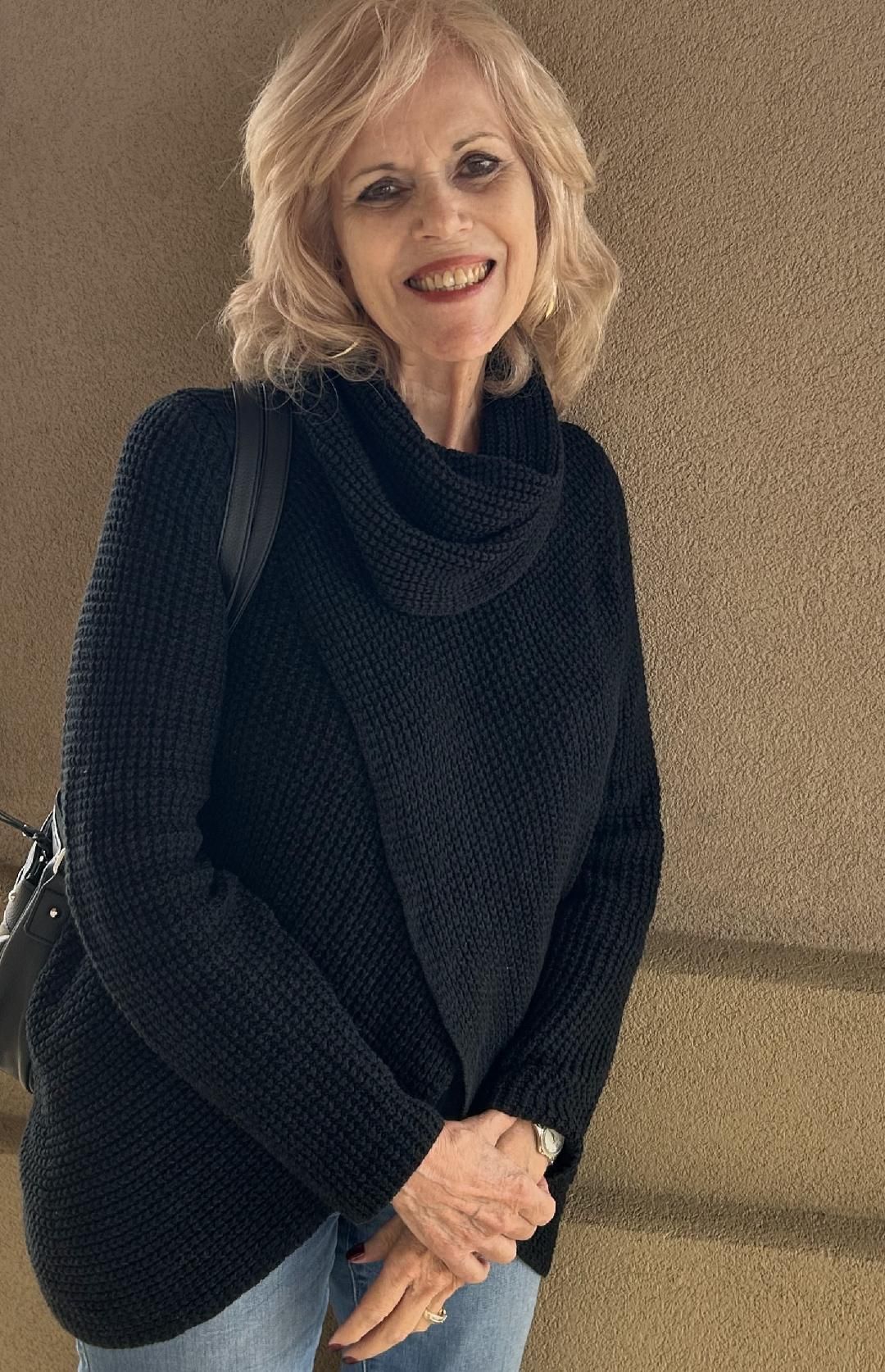 Smiling woman in a black sweater and jeans standing against a beige wall, holding a black shoulder bag.
