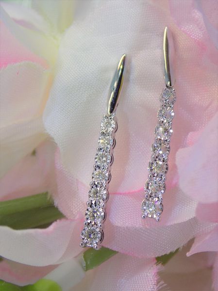 Diamond drop earrings with white metal settings, against a blurred pink background.