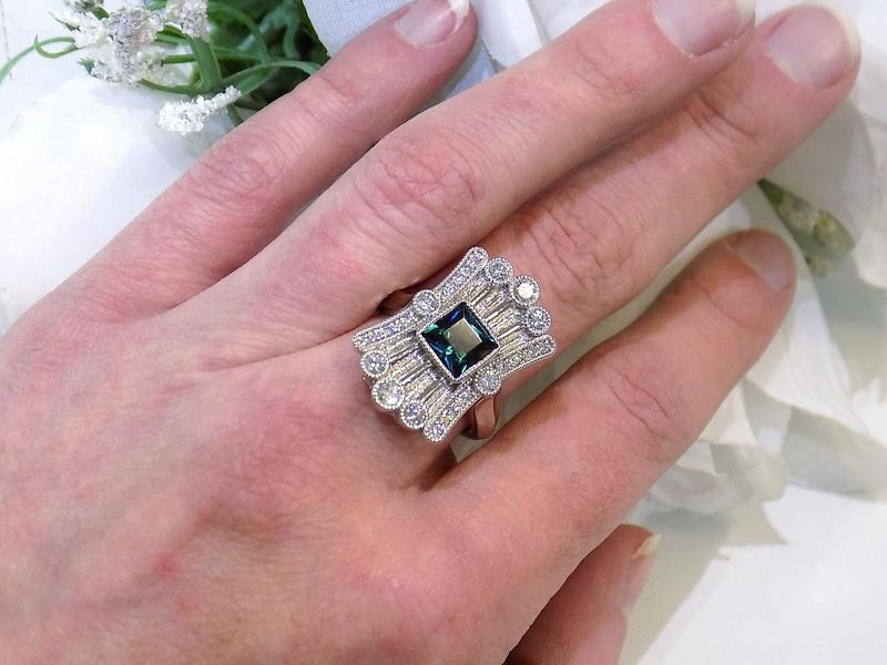 Hand wearing an ornate ring with a square teal gemstone and surrounding diamonds
