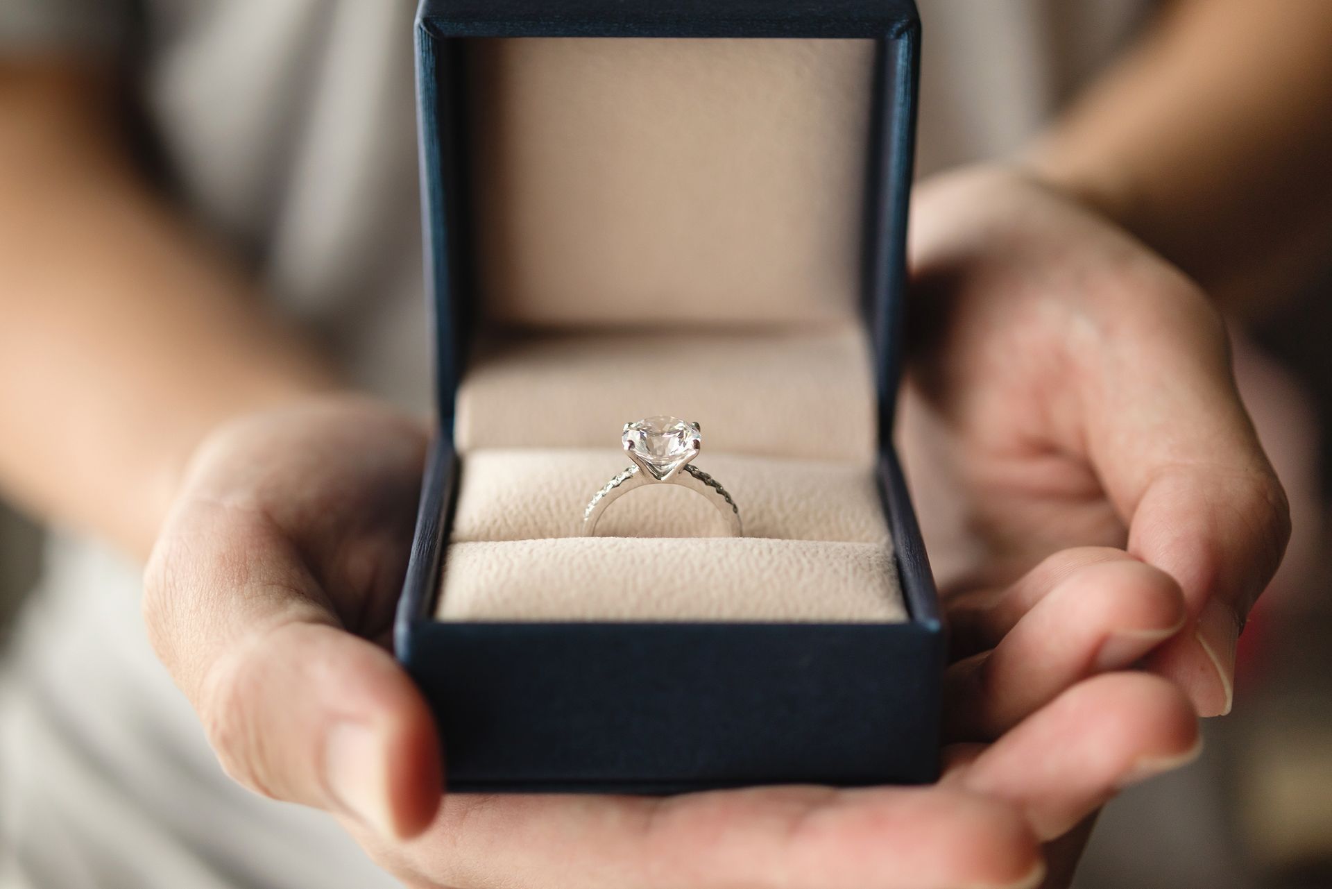 Hands holding a blue ring box open to display a diamond engagement ring