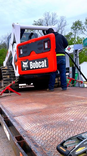 A person fueling a Bobcat skid steer on a flatbed trailer. Red and white machine.