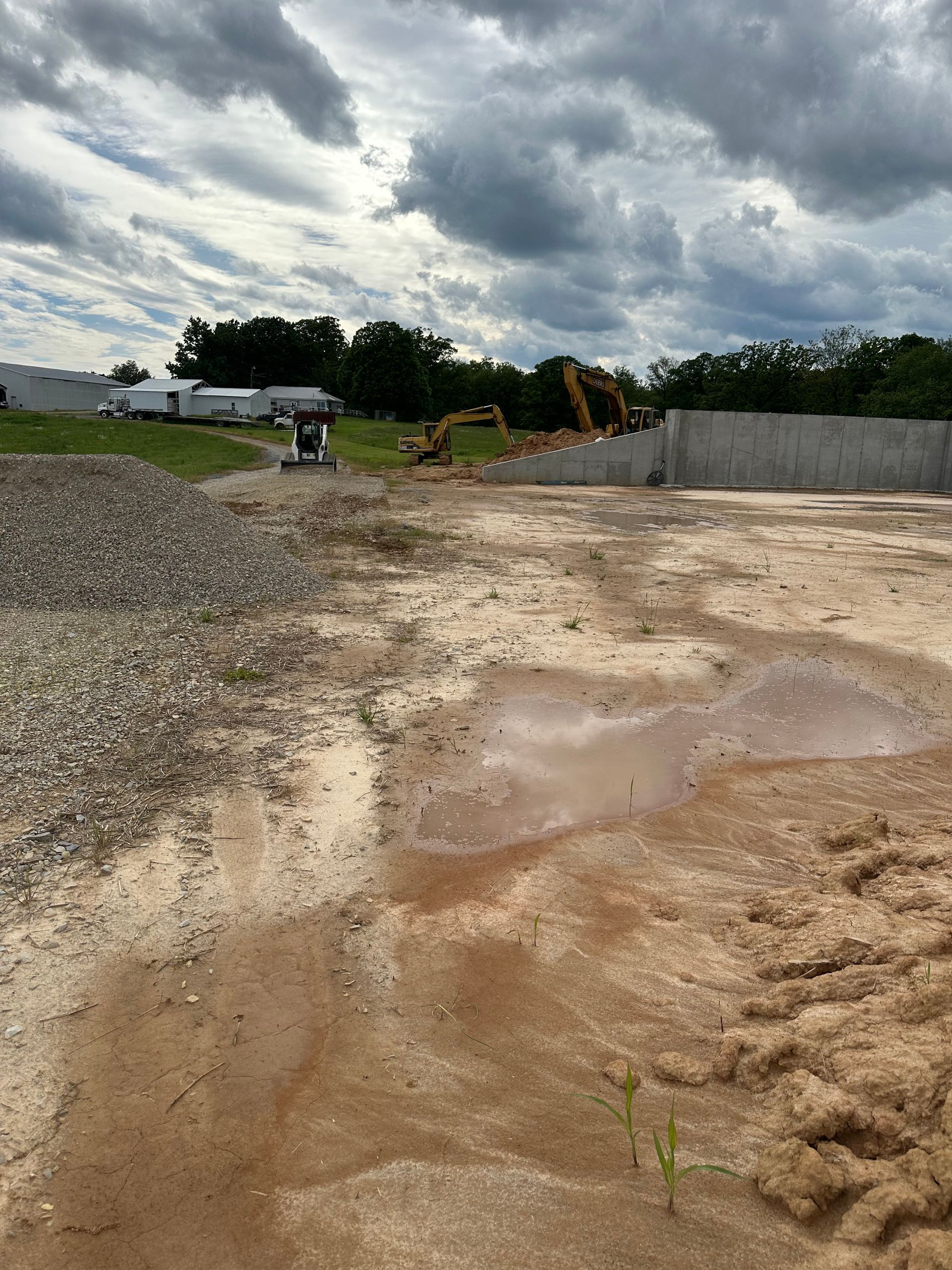 Construction site with gravel pile, puddle, and heavy machinery under cloudy sky.