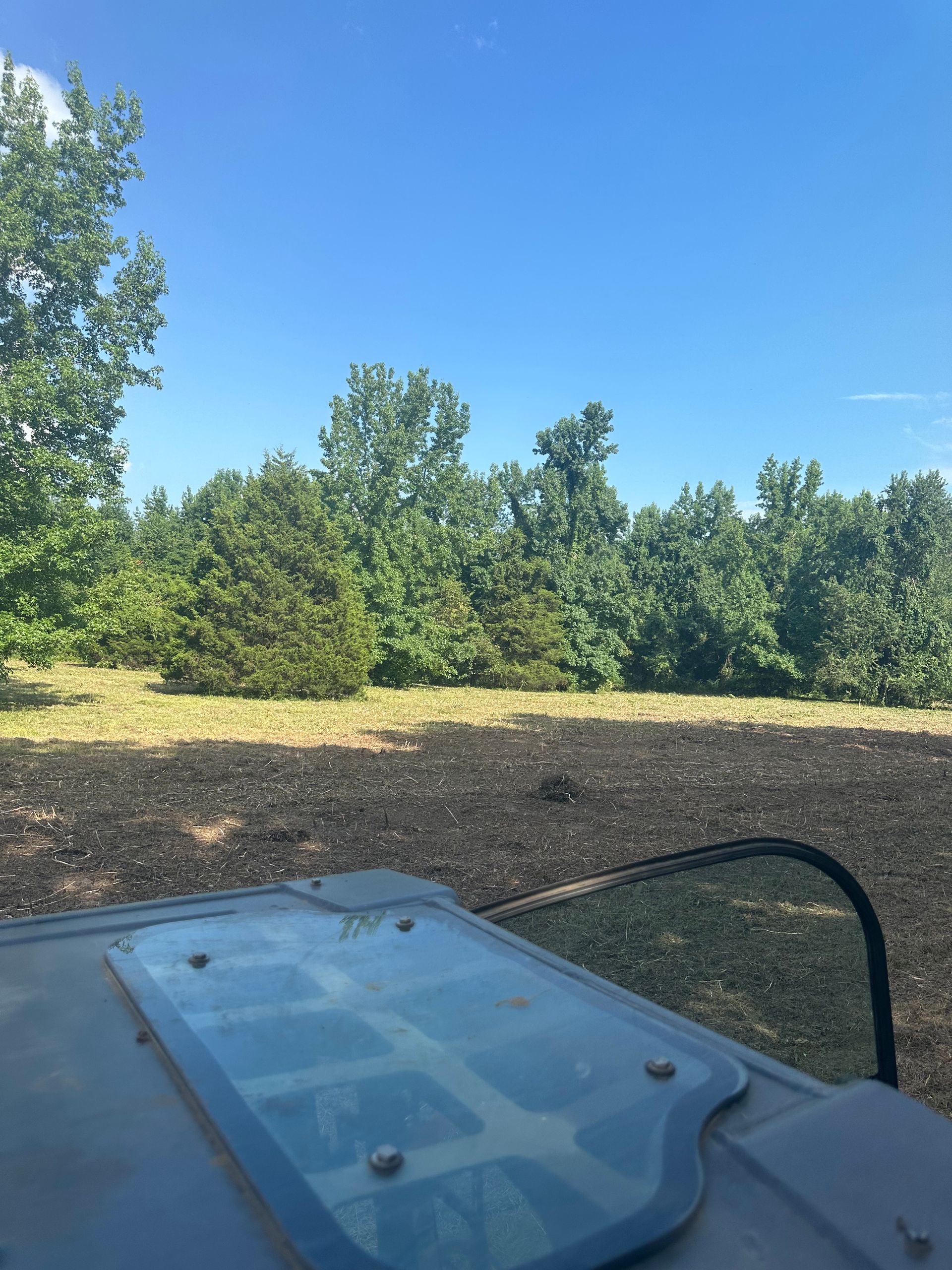 View from a tractor cab, looking at a freshly tilled field bordering a tree line, under a blue sky.