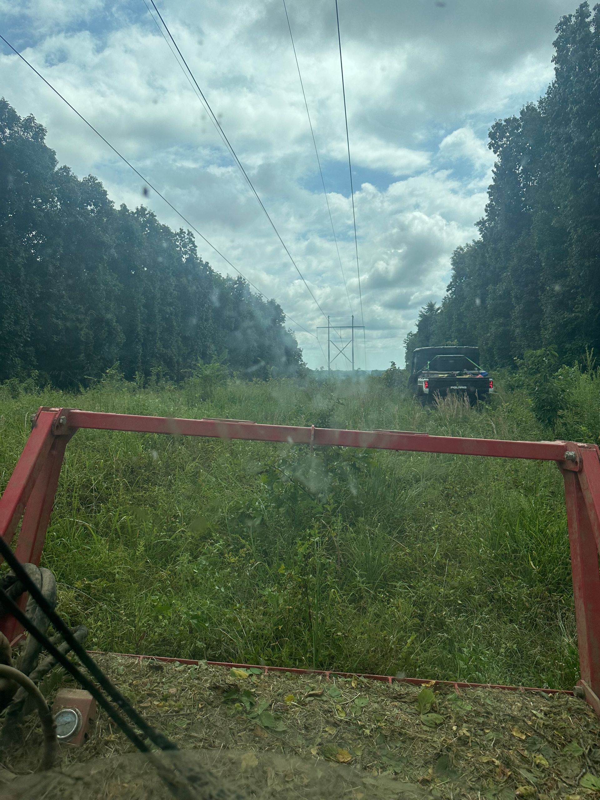 View from vehicle mowing a grassy path under power lines, trees on either side.