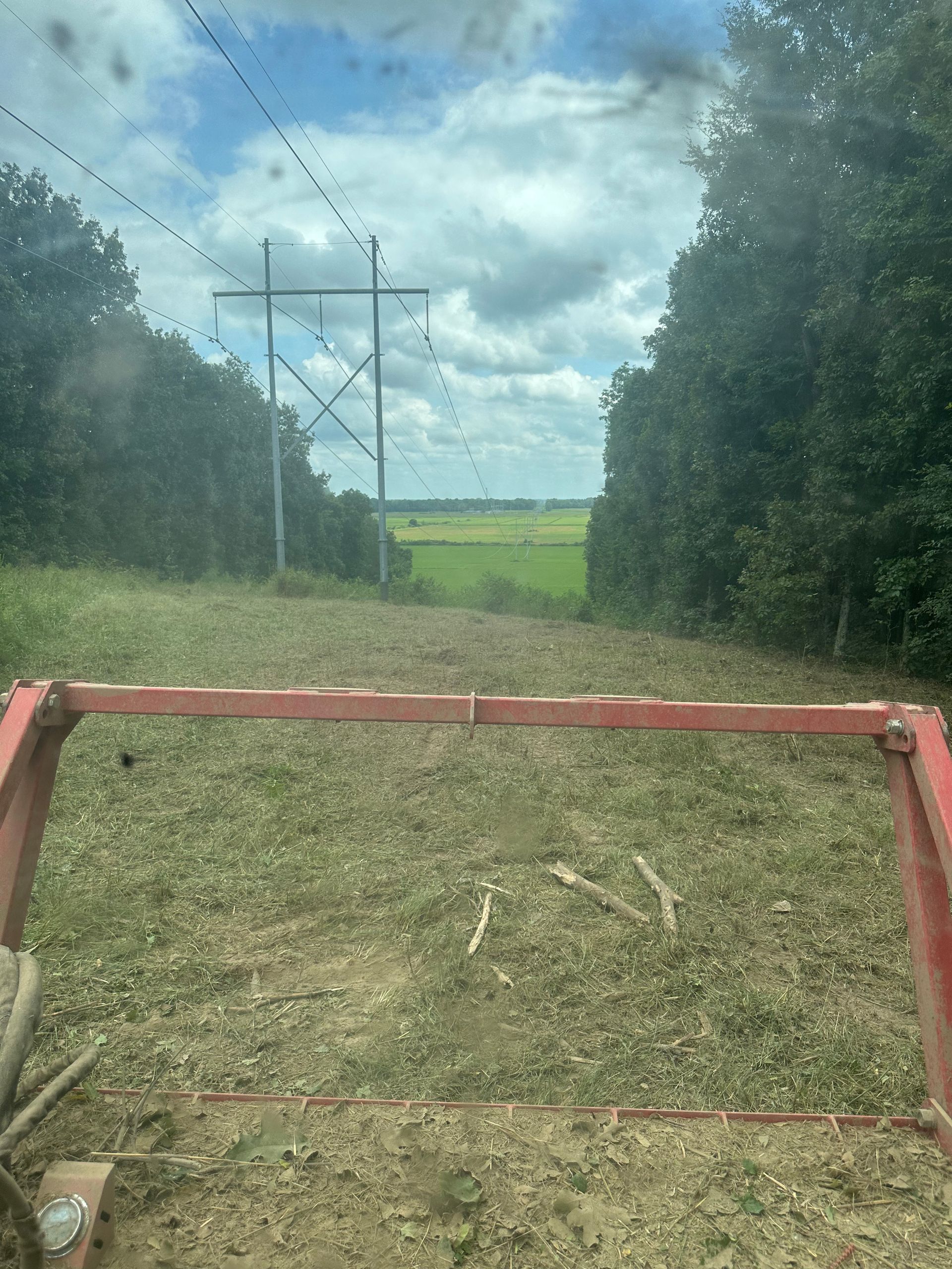 Red farm implement clearing a grassy path under power lines, flanked by trees, sunny day.