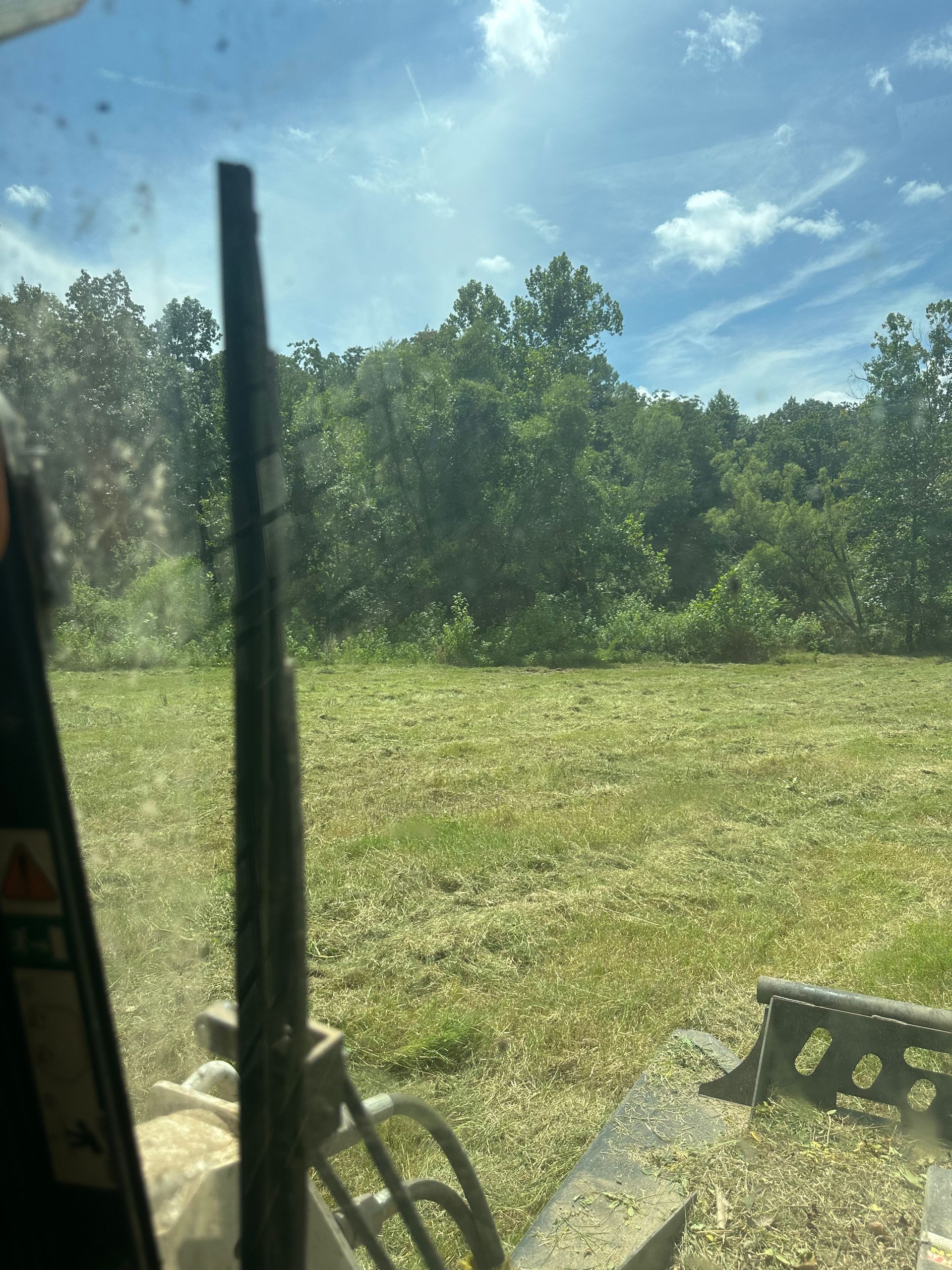 View from a vehicle's cab overlooking a field of mowed grass, trees, and a blue sky.