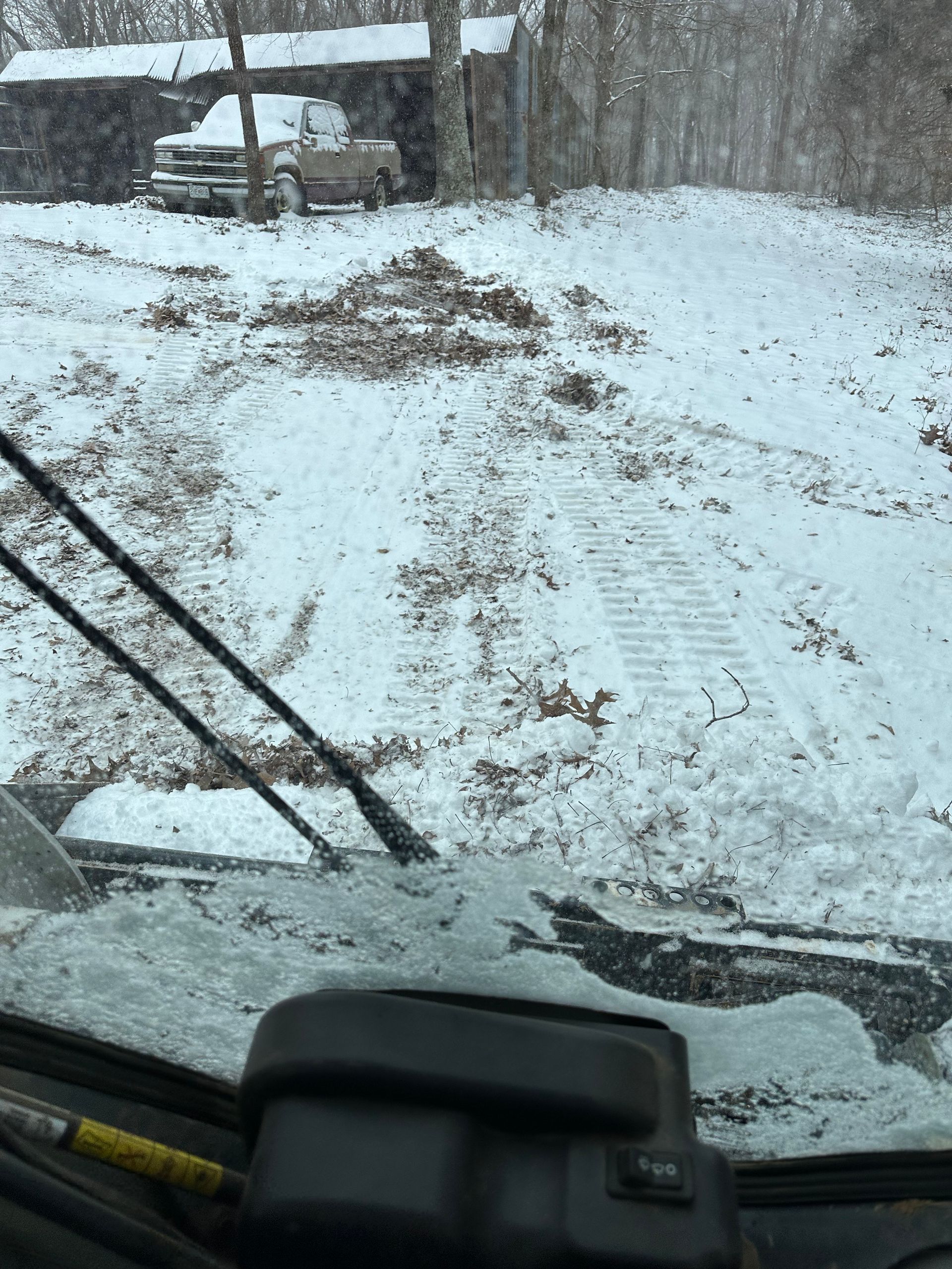 Snowy scene from a vehicle interior. A truck sits in front of a shed. Tracks in snow-covered dirt.