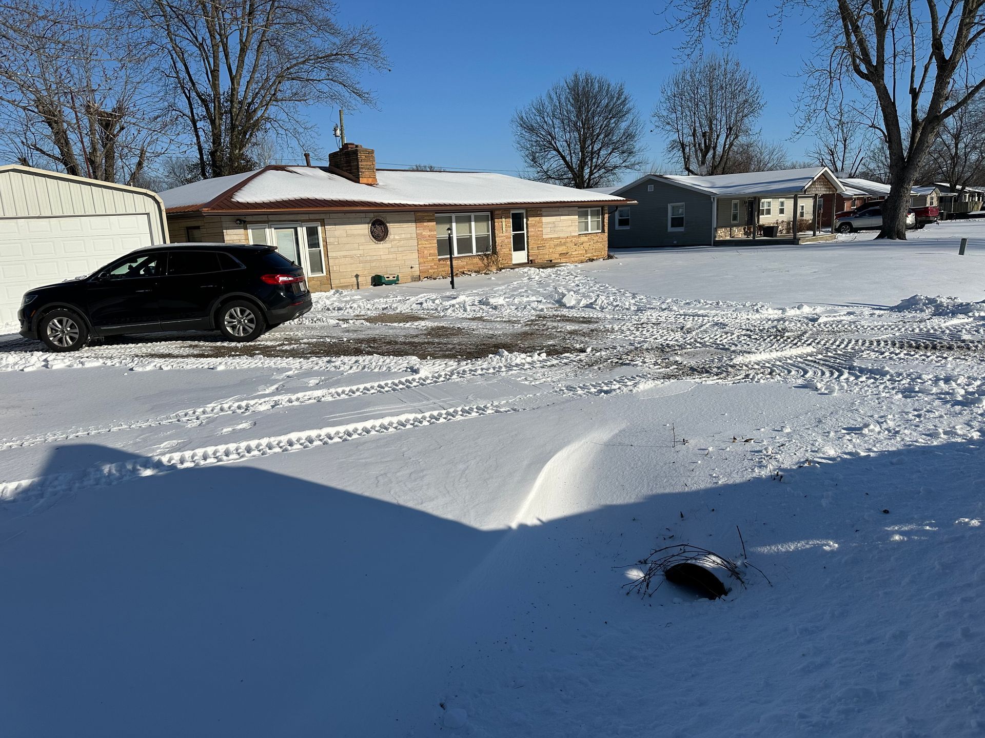 Snowy suburban scene; black SUV parked, houses in background, clear blue sky.