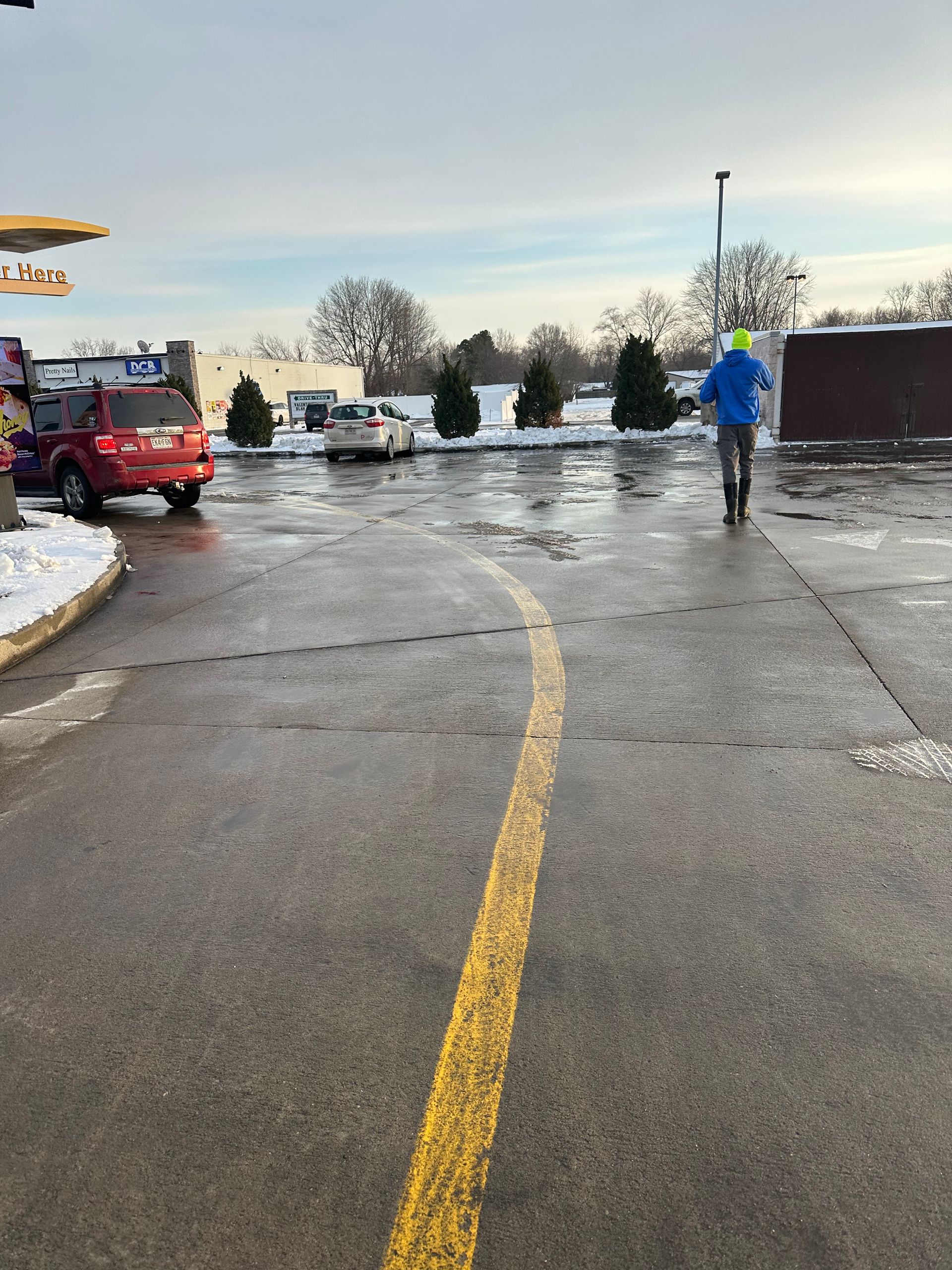 Person walking in a parking lot, yellow line in foreground, cars, snow, McDonald's in background.