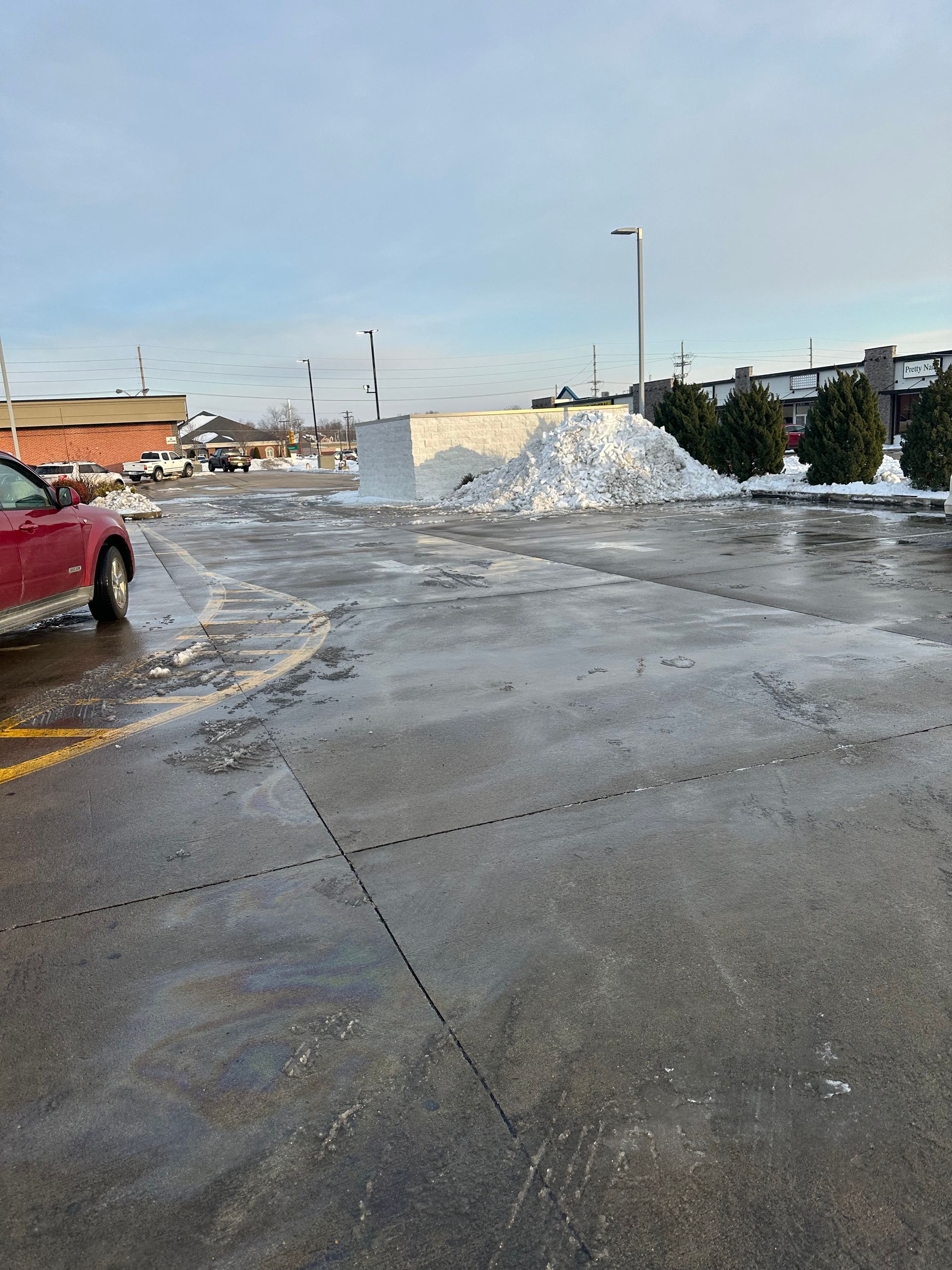 A snowy parking lot with a large snow pile. A red car is parked on the left.