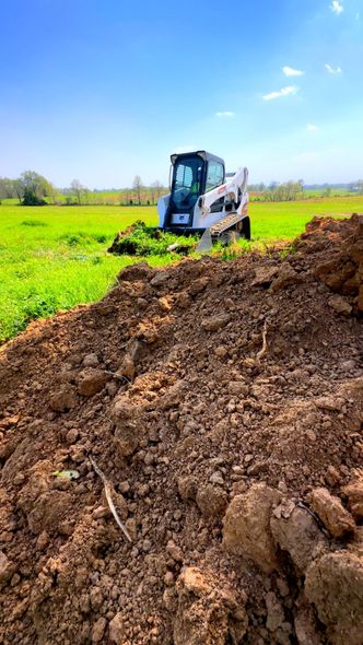 Bobcat skid-steer loader on a construction site, moving dirt in a green field under a blue sky.