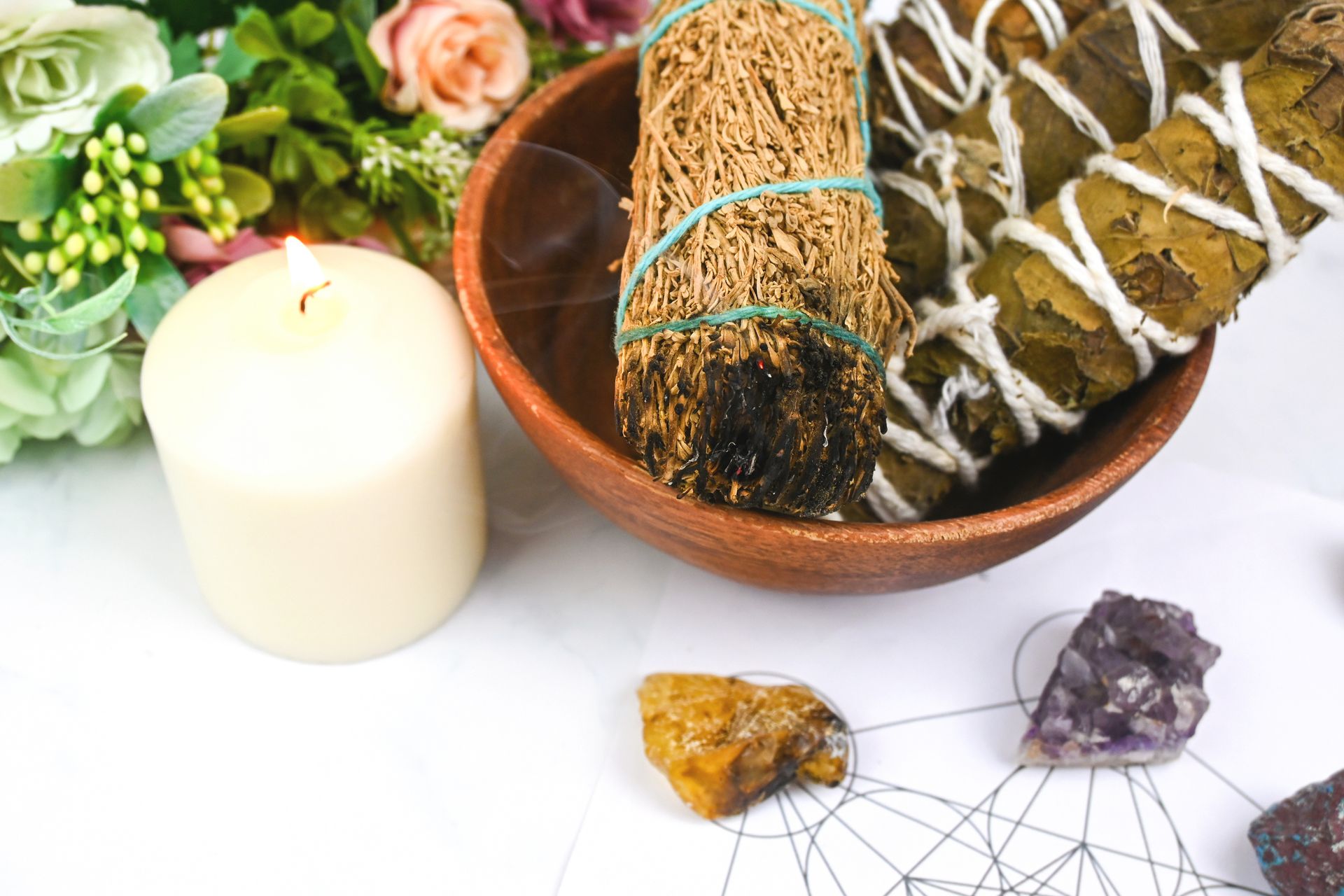 Candle, sage bundles in a clay bowl, and crystals arranged on a white surface with flowers nearby