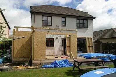 House under renovation with plywood extensions, blue tarps, and a picnic table in the yard