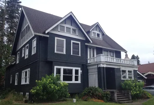 Large dark gray Victorian house with white trim, steep gables, and a front porch surrounded by greenery.