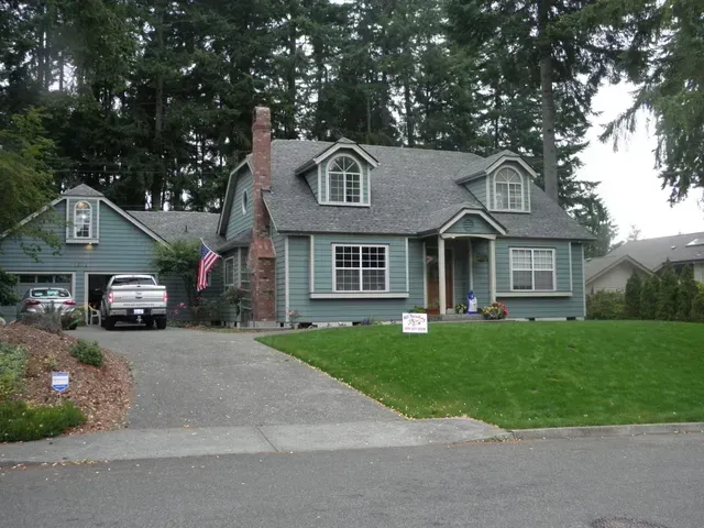 Blue house with green lawn, gravel driveway, parked SUV, and tall trees in the background