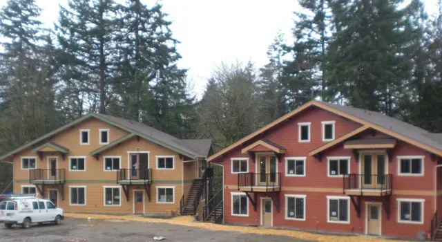 Two colorful apartment buildings in a wooded area, one tan and one red, with parked cars nearby