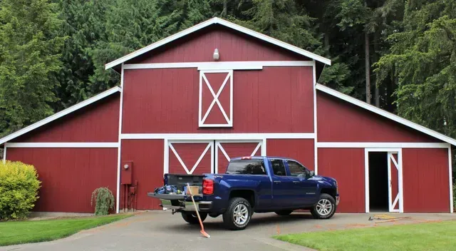 Red barn with white trim and a blue pickup truck parked in front on a grassy driveway.