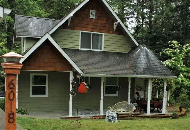 Small gray house with brown roof, front porch, and orange hanging decoration, surrounded by trees.