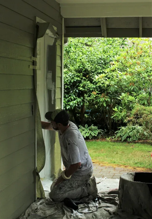 Painter kneeling on covered porch, painting the house wall light green near the doorway.