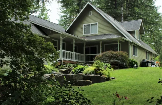 Gray house with a front porch on a lush green hillside surrounded by trees and shrubs