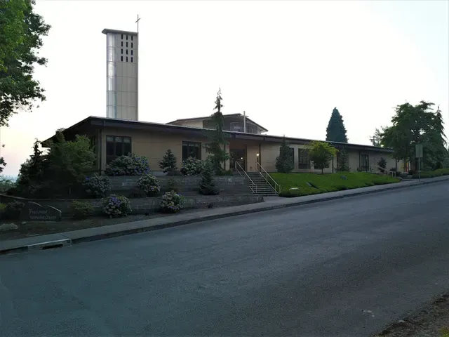 Church building with a tall white steeple beside a sloping road, surrounded by trees and shrubs.