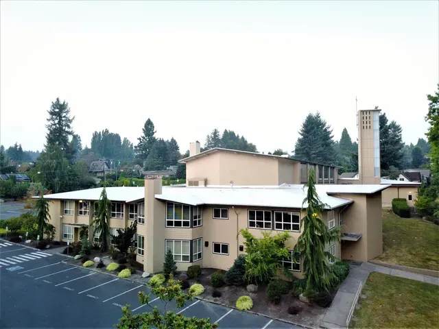 A beige multi-story building with trees and parking lot in front, seen from an elevated angle.
