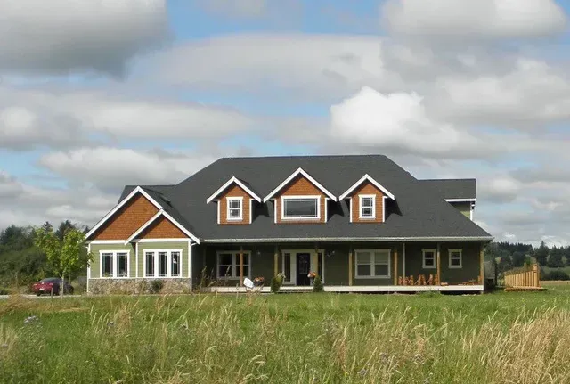 Large dark house with white trim and dormer windows in a grassy field under a cloudy sky