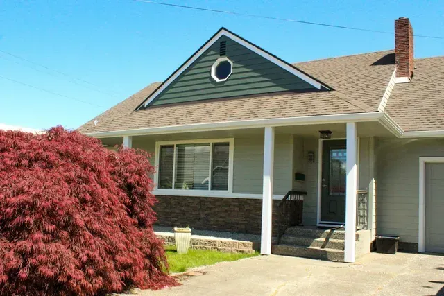 Single-story house with green siding, white porch, and red bush in front under a blue sky
