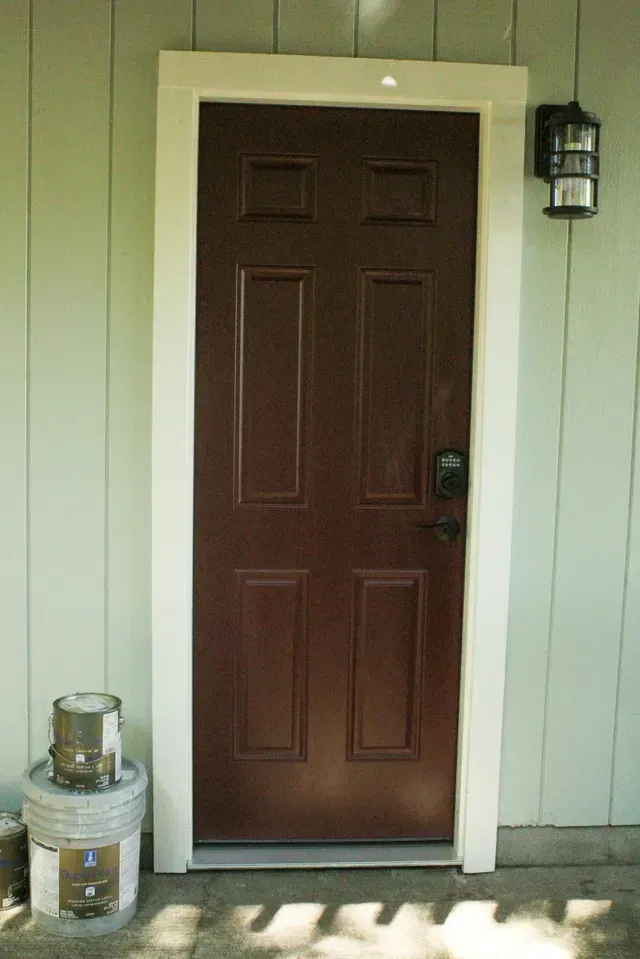 Brown front door with white trim on a beige house exterior, with a lantern light and small stool beside it