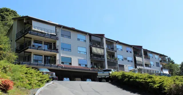 Modern apartment building on a hillside with balconies, green shrubs, and a clear blue sky