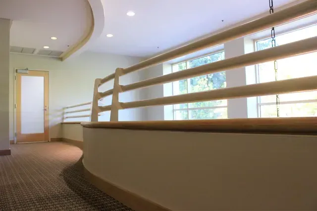 Curved indoor hallway with carpet, white railings, and a window view of trees