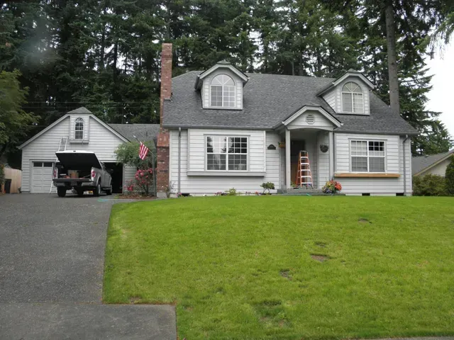 White house with detached garage, driveway, and green lawn in a wooded yard