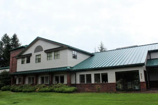 Large farmhouse with green metal roof and brick porch, set on a grassy lawn under an overcast sky