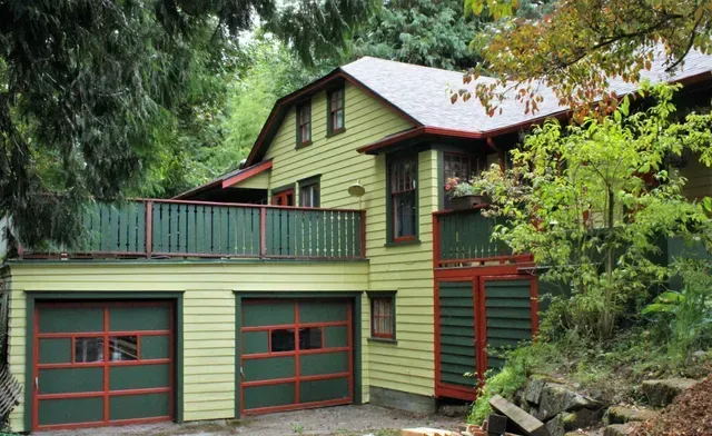 Green house with red trim, double garage doors, and a balcony surrounded by trees.