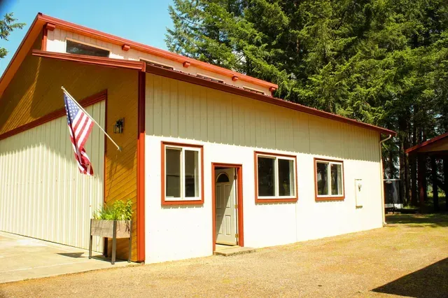 Small cream-colored house with red trim and an American flag outside in a wooded area