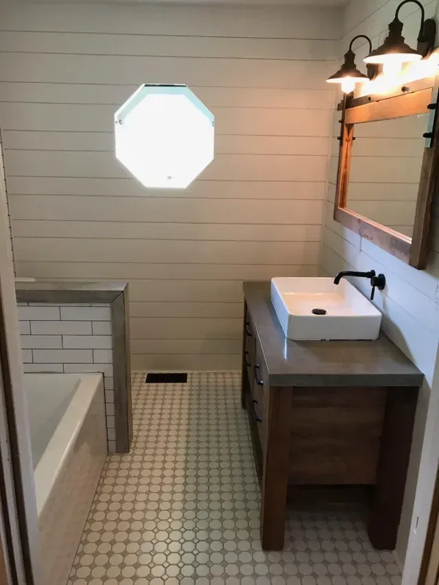 Small bathroom with bathtub, vanity sink, mirror, and octagonal window over patterned tile floor.