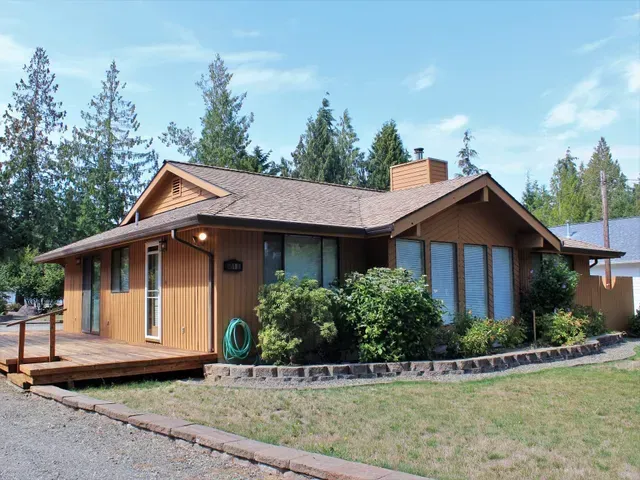 Wooden cabin with a brown roof, green bushes, and tall pine trees under a blue sky