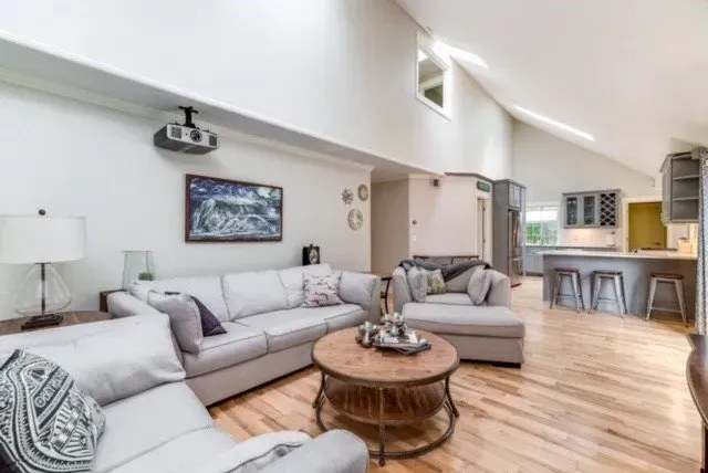 Bright open-concept living room with gray sofas, round wooden coffee table, and kitchen bar stools in the background