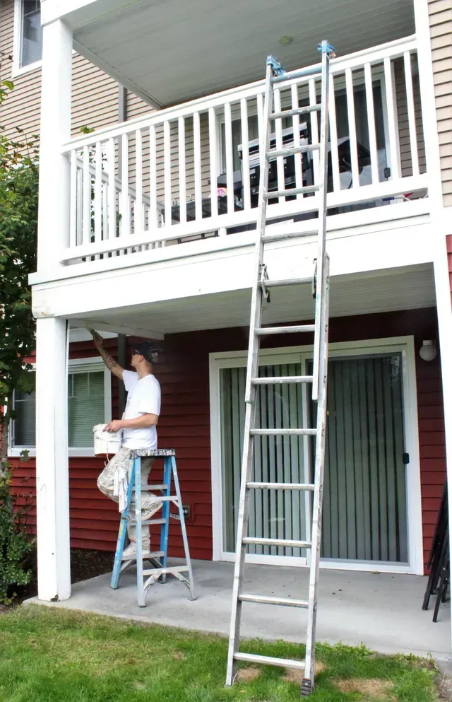 Person on step stool cleaning a second-story balcony with a ladder leaning nearby