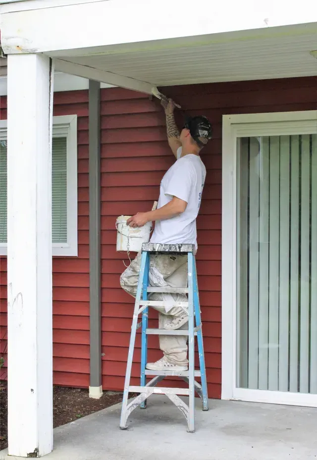 Person standing on a ladder painting the ceiling of a red house porch