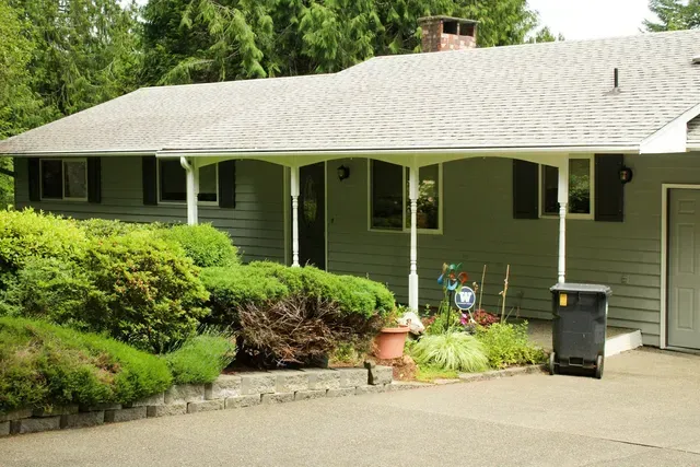 Green house with a covered porch, shrubs, potted plants, and a driveway in front.