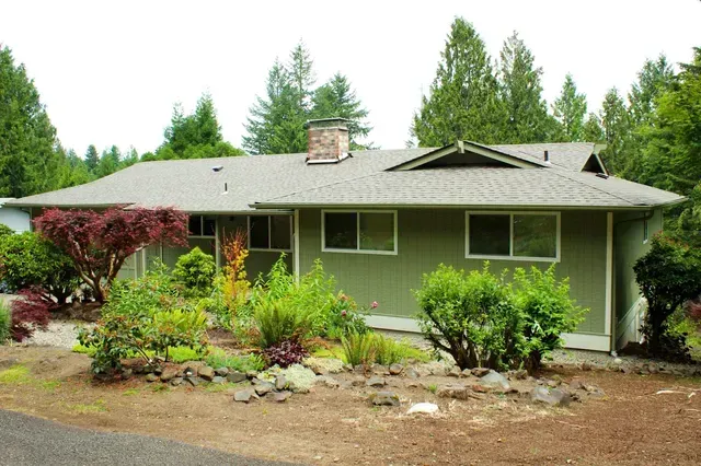 Single-story green house with a landscaped front yard and trees in the background