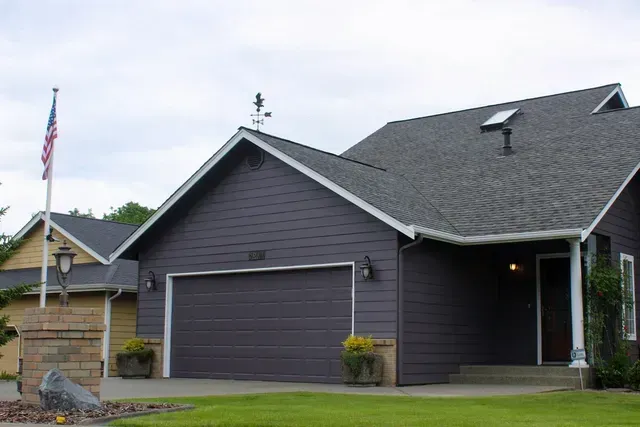 Suburban house with dark gray siding, attached garage, green lawn, and American flag on a cloudy day