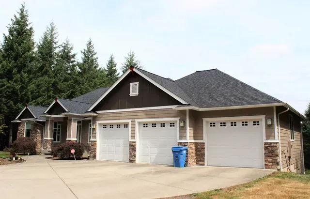 Three-car garage with beige siding and dark roof, set beside tall trees and a driveway