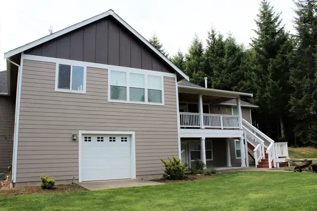 Two-story beige house with garage, white trim, and a deck beside tall evergreen trees.