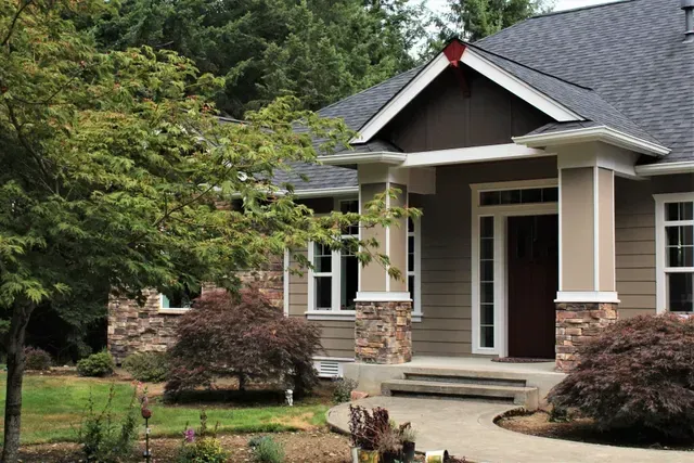 Small beige house with stone accents, front porch, and landscaped yard surrounded by trees.
